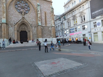 Kolejny przedziwny kadr, w centrum Sarajewa: katedra, przed nią Róża Sarajeva (ubytek wypełniony czerwoną farbą, miejsca, gdzie wybuchały bomby śmiertelnie raniąc ludzi), bawiące się dzieci, kredowy malunek i poster o wystawie „Srebrenica”. W żadnym innym